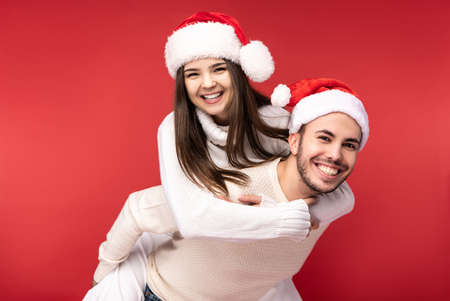Photo of sweet couple in Christmas hats, happy about holidays, she jumped on his back. Male and female are in love, look blessed and smile, isolated over red backgroundの写真素材