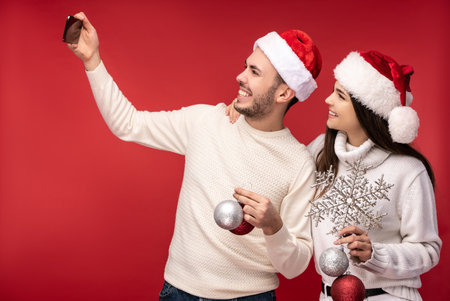Photo of sweet couple in Christmas hats and with Christmas toys, looks happy. Male and female take a selfie and smile, isolated over red backgroundの写真素材