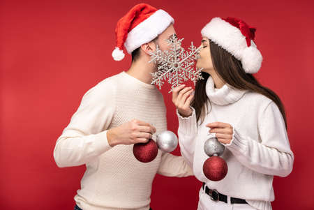 Photo of sweet couple in Christmas hats and with Christmas toys, kiss each other. Male and female are in love look blessed and smile, isolated over red backgroundの写真素材