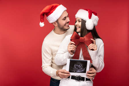 Photo of sweet couple in Christmas hats, happy about having baby. Male and female are in love look blessed and smile, isolated over red backgroundの写真素材
