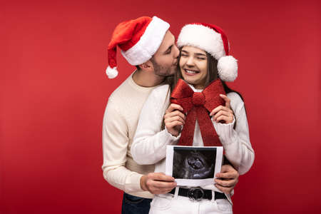 Photo of sweet couple in Christmas hats, happy about having baby. Male and female are in love look blessed and smile, isolated over pink backgroundの写真素材