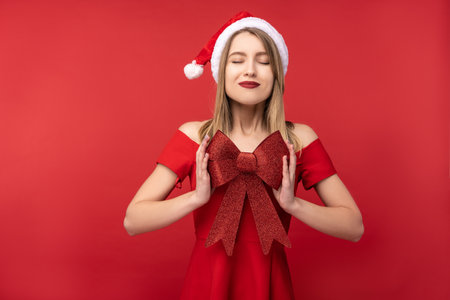 Photo of attractive woman in Christmas hat and red dress, holds red bow for present and make a wish, Isolated over red backgroundの写真素材