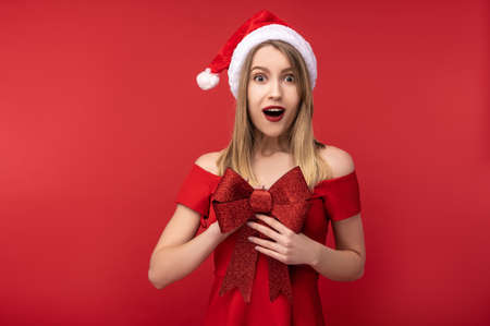 Photo of attractive woman in Christmas hat and red dress, holds red bow for present and look with surprise, Isolated over red backgroundの写真素材