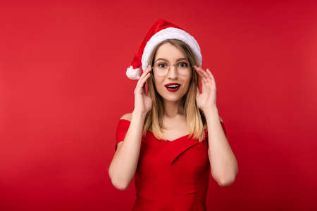 Photo of attractive woman in Christmas hat and red dress, holds glasses with surprised. Isolated over red backgroundの写真素材