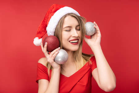Photo of attractive woman in Christmas hat and red dress, plays with Christmas toys and poses with smile. Isolated over red backgroundの写真素材