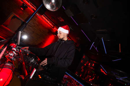 New Year in night club. Merry Christmas Young confident DJ in red santa claus hat mixing music on turntables at holiday party. Shining disco ball. Black and red club background.の写真素材