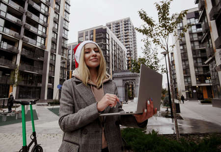 Blonde woman in santas hat wearing casual clothes sitting outside and working on her laptop. Merry Christmas and Happy New Year Young freelancer woman doing her work on fresh air.の写真素材