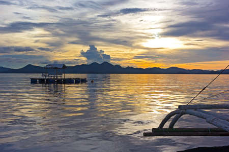 A view from traditional phishing boat with outriggers in lagoon of Coron Island at cloudy sunset. Floating gazebo at sunset with mountain ridges on horizon.の写真素材