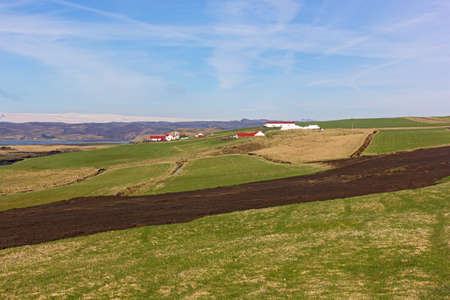 Agricultural landscape in Iceland. Scenic fields with farm houses, volcanic formations and mountains with snow peaks on horizon.の写真素材