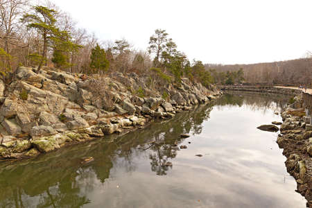 Rocky banks of the canal along Potomac River with footpath. Winter forest landscape with trees reflection in canal waters.の写真素材