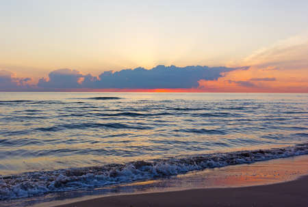 Cloudy sunrise and sea waves rolling on an empty sandy beach. Sun appears above clouds during dawn over the Mediterranean Sea in Valencia region of Spain.の写真素材