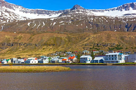 Landscape of coastal town in Northern Iceland during spring. Mountain tops covered in snow, while waterfalls located beneath them rushing towards the sea waters.の写真素材