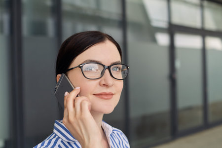 Business woman with brown hair in glasses in a blue and white striped shirt with white cuffs on her...