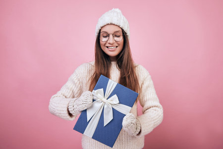 Cheerful happy girl is standing in pink back in glasses, milk-colored mittens and a hat, in her...
