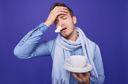 An sick guy in a denim shirt and a blue scarf around his throat is standing with a napkin in his...