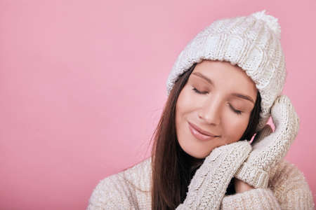 Cute girl with closed eyes in a knitted milky gray sweater and hat stands on the background. The atmosphere is cozy, warm in cold weather. As if sleeping on her hands in mittens.の写真素材