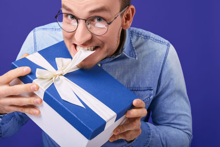 Cheerful young man in a blue denim shirt and round glasses standing with a gift with a blue cap in his hands, trying to untie him with his teeth. Xmas, New Year, Valentine's Day.の写真素材