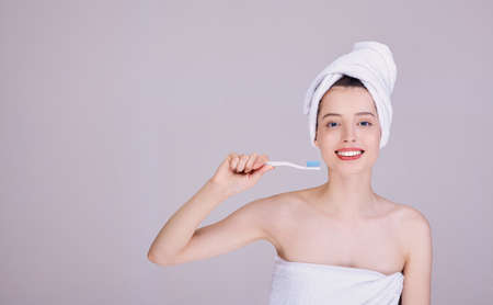 A beautiful half-naked girl in a towel, after a shower, holds a toothbrush with blue bristles, conducts a morning routine. Dental care and dental health.の写真素材
