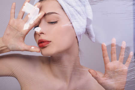 Young beautiful girl after a shower, with a towel on her head, tries to break through the cellophane. Looking away. Silicone cosmetics. The concept of health and beauty. Organic cosmetics.の写真素材