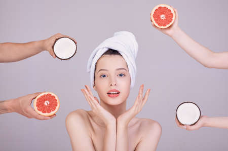 Surprised girl with beautiful skin, in a white towel on her hair, half-naked, holds her hands by her face. Around the girl, several pairs of hands hold coconuts and grapefruits.の写真素材
