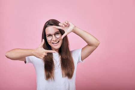 Cute happy cheerful girl in round glasses standing on a pink wall back and smiling slightly. Life style, emotions. Woman shows frame borders for photo with her hands. Copy space, close up.の写真素材