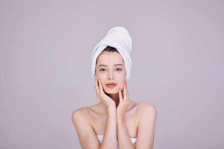 Image of an adorable half-naked woman wrapped in a towel, smiling after a shower with hands on her face. Sensuality and tenderness. Care and care. Beauty concept. Natural beauty. Face and body care.の写真素材