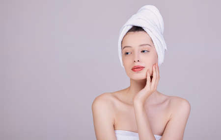 A pensive woman with dark hair, with a towel on her head, after a shower, touches her face with her left hand. Sensuality and tenderness. Care and care. Beauty concept. Face and body care.の写真素材