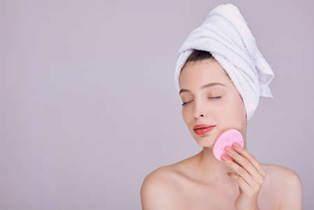 Gorgeous girl with nude make up and naked shoulders posing on gray background with cleaning sponge, skin care concept, beauty photo. Caring and youthful skin. Body care.の写真素材