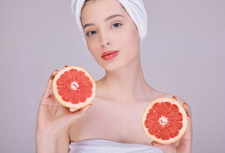 Portrait of a young beautiful woman with healthy glow of perfectly smooth skin holds two slices of grapefruit. Natural cosmetics, skin care. Half-naked, in a white towel on her head, looks straight.の写真素材