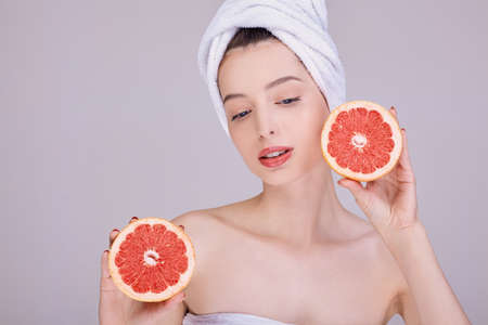 Half-naked young attractive girl with clean skin, in a towel on her head, holds a freshly cut grapefruit in her hands near her face. A look to the side. Beauty Porter. Photo on the background.の写真素材