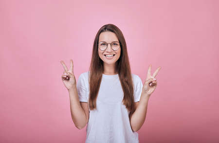 Cheerful cool girl in a white T-shirt and round glasses standing on a pink background and smiling. Life style, emotions, happiness. Woman shows fingers peace.の写真素材