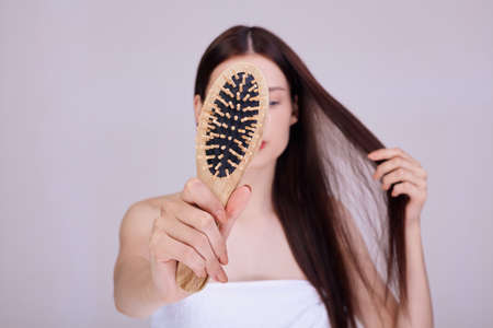A cute brunette wrapped in a white bath towel shows a wooden comb right in the frame, covers her face. Hair care, hydration and volume for hair. Hair loss.の写真素材