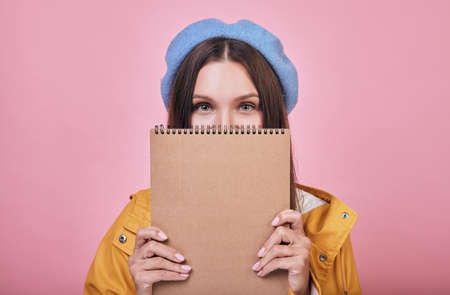 Cool co-ed with beaytiful green eyes and dark hair in blue headdress and yellow raincoat is standing with a brown notebook in her hands. Lady looking straight open.の写真素材