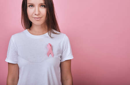 Cute charming girl in a white T-shirt with long dark hair smiling slightly. Emotions. A woman is standing calmly, with a pink ribbon on her chest.の写真素材