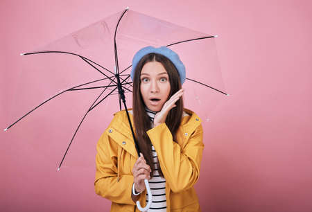 Pretty surprised girl in a gentle blue barret, a striped blouse and a yellow rain jacket smiles cute against a pink background with umbrella in hand. Arm near her face, looking forward with open mouthの写真素材