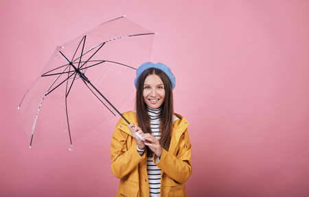 Pretty happy joyful girl in gentle blue barret, striped blouse and a yellow rain jacket against pink wall back, umbrella in both hands. Lady looking straight and smiling. Rainy autumn weatherの写真素材
