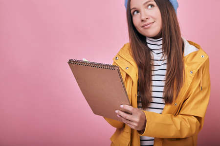 A cute educated girl with a charming smile in a yellow jacket, a striped sweater and a blue headdress is standing with a notebook and pen in her hands thoughtfully. Education, writing, composition.の写真素材