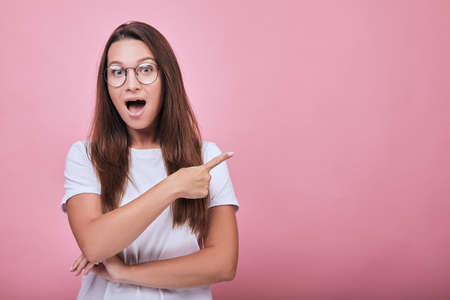 Cute surprised woman in glasses with dark hair standing on a pink background. Lady shows forefingers to the side and her mouse is open. Life style, emotional.の写真素材