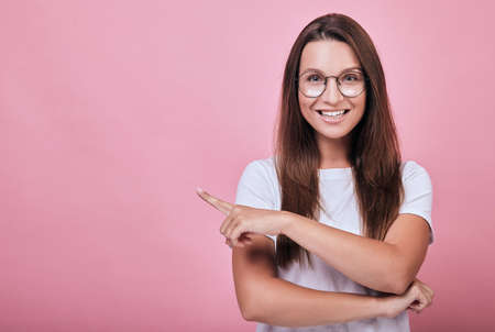 Attractive joyful cool girl in a white T-shirt with her hair standing on a pink background and smiling broadly. Life style, emotional. Lady shows forefingers to the side.の写真素材