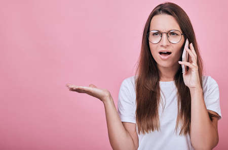 Joyful funny charming girl in a white T-shirt with her hair on a pink background and smiling slightly. Emotional, speaking. Woman talking on a mobile cell phone in surprise.の写真素材