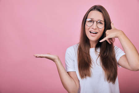 Cute funny charming girl in a white T-shirt standing on a pink background. Life style, emotions. The woman folded the fingers of one hand as if talking on the phone.の写真素材
