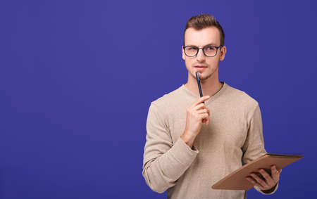 Young confident guy redactor, a professional in his field. A pensive man stands on a purple background in a beige sweater with a brown paper notebook in hand and black ballpoint pen near his face.の写真素材