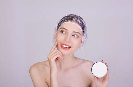 A smiling brunette in a shower cap holds a cream in her hand and puts it on her face with a finger. Playfully looks away. Face cream. Pprocedure for the beauty of the body. Caring for yourself.の写真素材