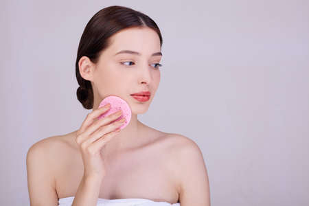 Attractive woman with dark tied hair holds a makeup sponge on her face, takes care of her face, removes pores, poses half naked on the light background.の写真素材