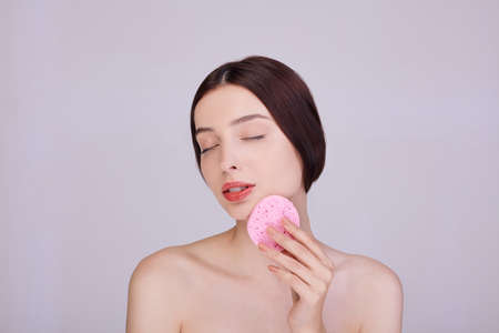 A beautiful woman with dark hair holds a makeup sponge on her face, takes care of her face, removes pores, poses half naked, radiates calm. Tenderness and care. Personal care.の写真素材