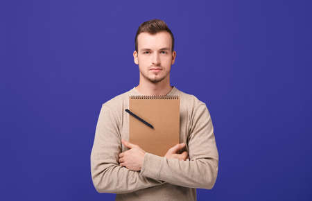 A young confident confident guy. The creator calmly looks forward. The writer stands on a purple background in a beige sweater, hugging a brown notebook with black ballpoint pen in his hands.の写真素材