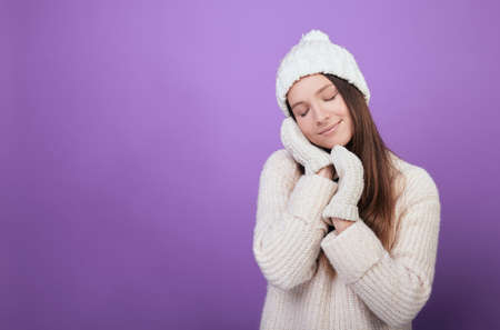 The girl closed her eyes and folded her hands in mittens under her cheek. A young brown-haired woman in a good mood in a warm white sweater and white mittens. Photo on a purple background.の写真素材