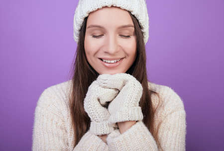 Cute girl in a white knitted hat and white mittens folded her hands under her chin. Looks down, embarrassed, smiling. Photo on a purple background. The atmosphere of comfort.の写真素材
