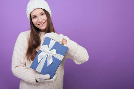 A young brown-haired woman in a white knitted sweater, hat and mittens holds a gift in her hands with a blue cap and white ribbon. He looks straight and smiles gently. Christmas spirit.の写真素材