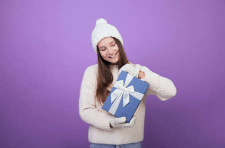 A young woman in a white knitted sweater, hat and mittens holds a gift in her hands with a blue cap and white ribbon. Looking for a present and smiles. Good New Year spirit.. Christmas presents.の写真素材
